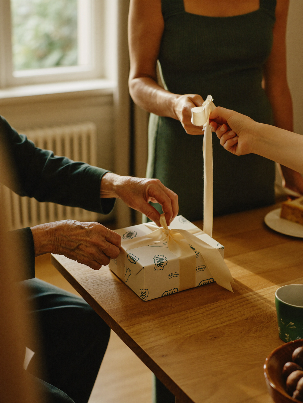 Hände von Großmutter und Enkelin öffnen gemeinsam ein Geschenk am Küchentisch